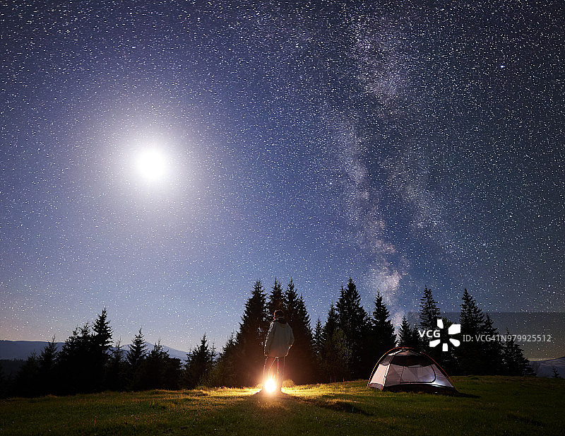 男徒步者享受夜晚露营附近的旅游帐篷在篝火下蓝色星空和银河图片素材