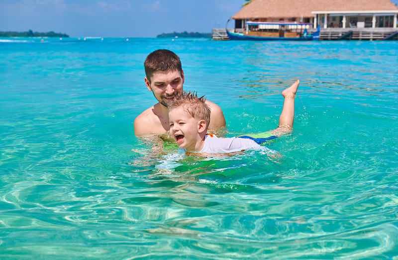 三岁蹒跚学步的小男孩跟着爸爸学游泳。暑假全家去马尔代夫度假。图片素材