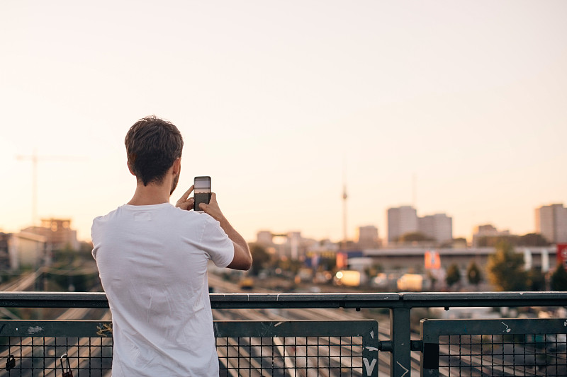 在晴朗的天空下，站在桥上用手机拍摄城市的年轻人的后视图图片素材