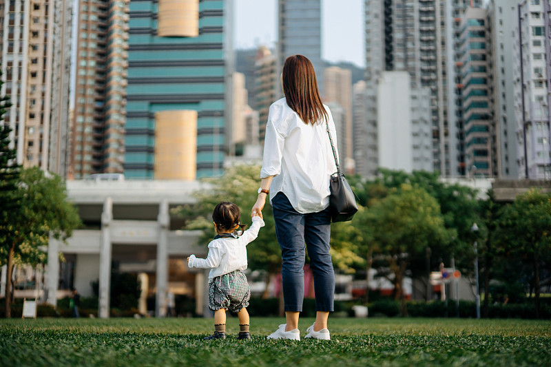 后视图年轻的母亲和女儿手牵手走在城市公园对现代城市天际线图片下载