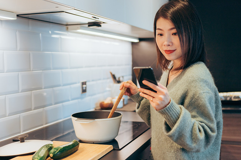 年轻开朗漂亮的女人在家里做饭的时候在智能手机上阅读食谱图片素材
