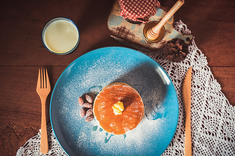 木桌背景上的早餐是蓝色盘子里的牛奶、蜂蜜和杏仁煎饼。图片下载