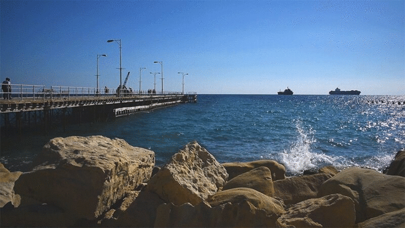 塞浦路斯利马索尔，海浪拍打着码头附近的岩石，背景是地平线上的两艘船图片下载