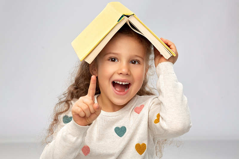 阅读，教育和童年的概念-肖像的微笑小女孩与书在屋顶上在灰色的背景。头戴书微笑女孩的肖像图片下载