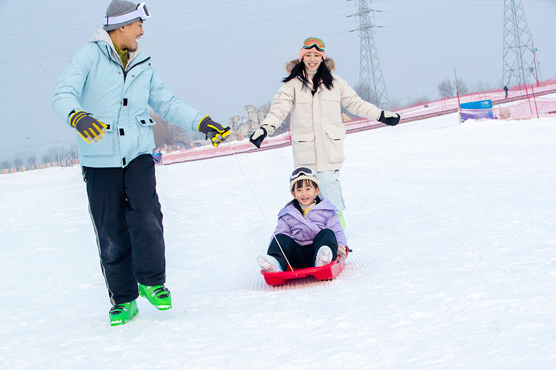 滑雪场上父母和坐在雪上滑板的女儿玩耍图片下载
