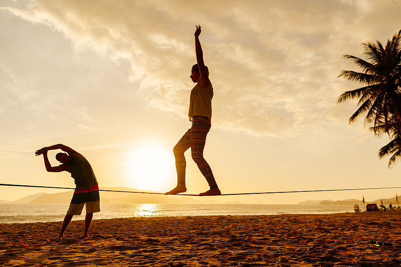 侧面的十几岁的女孩在钢丝上平衡与前面的男人伸展在海滩上的观点图片素材