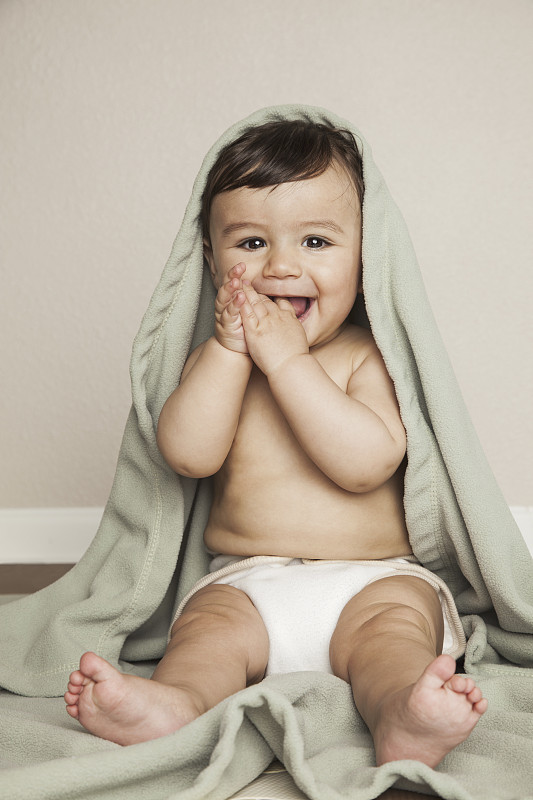 一个8个月大的小男孩穿着布尿布，坐在地板上。他头上盖着一张帆布床。图片下载