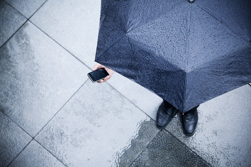 高视角的商人拿着伞，看着他的手机在雨中图片素材