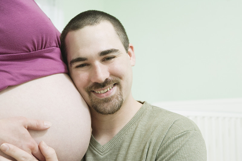 男人在育儿室拥抱孕妇，耳朵贴着孕妇的肚子，微笑着图片下载