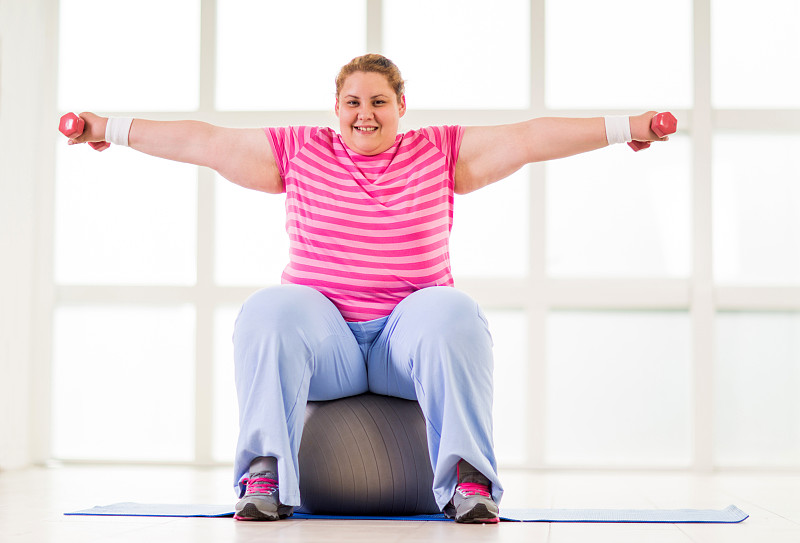 微笑的胖女人坐在健身球和举重。图片下载