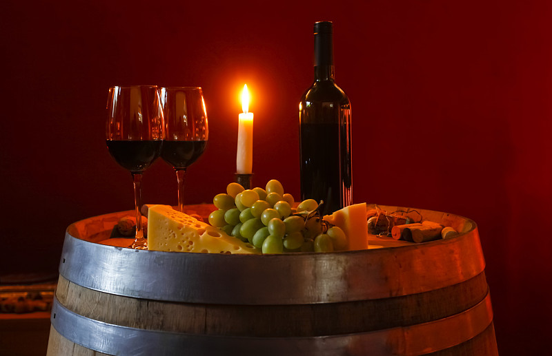Two glasses of red wine, bottle, grapes, cheese and lighted candle on a wine cask in a wine cellar图片素材