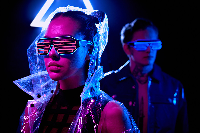 年轻的网络女人的肖像在未来的眼镜和透明夹克在前景，男人在霓虹灯眼镜站在背景图片素材