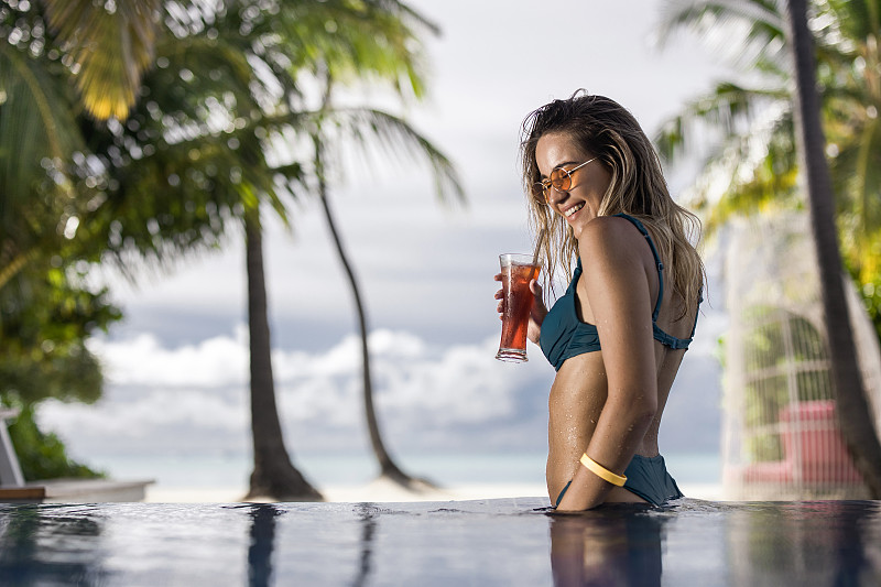 年轻快乐的女人在海滩上的游泳池里喝着鸡尾酒。图片下载