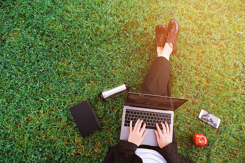 公园里的一个女孩坐在草地上用笔记本电脑工作，一个女人的手放在键盘上。靠近手机，苹果，笔记本。学习在户外。远程学习的概念图片素材
