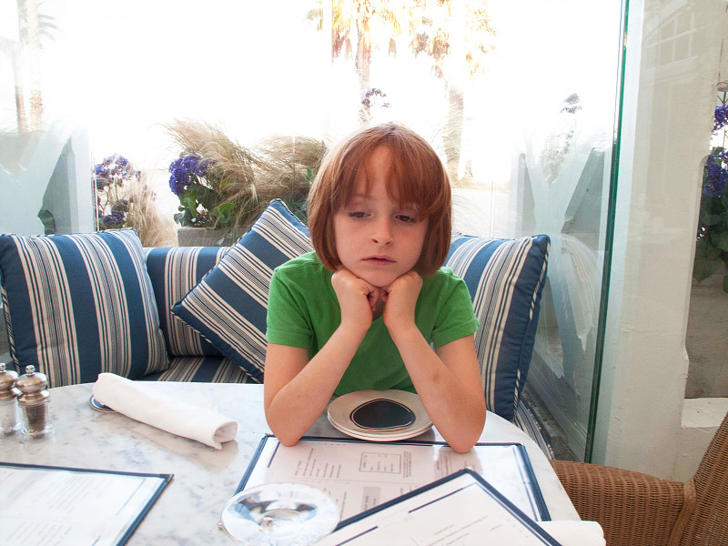 一个红发小女孩独自呆在咖啡馆窗前，看起来很无聊。图片素材