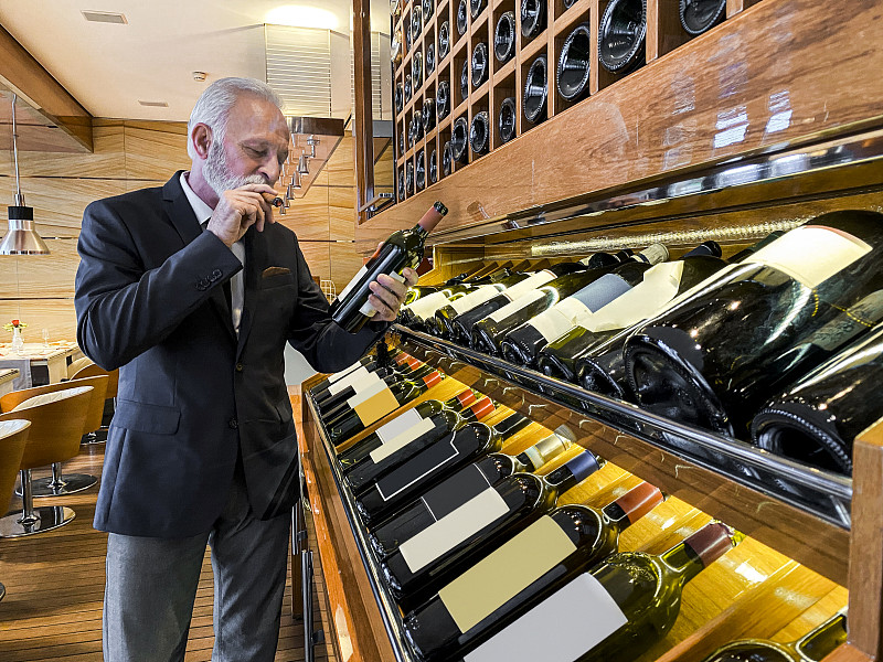经验丰富的年长侍酒师选择一个葡萄酒品尝在一个豪华的酒窖。他在抽雪茄图片素材
