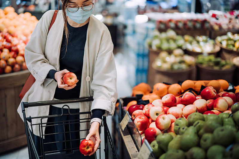 2019冠状病毒病健康危机爆发期间，年轻的亚洲女性戴着防护口罩在超市购买新鲜食品图片素材