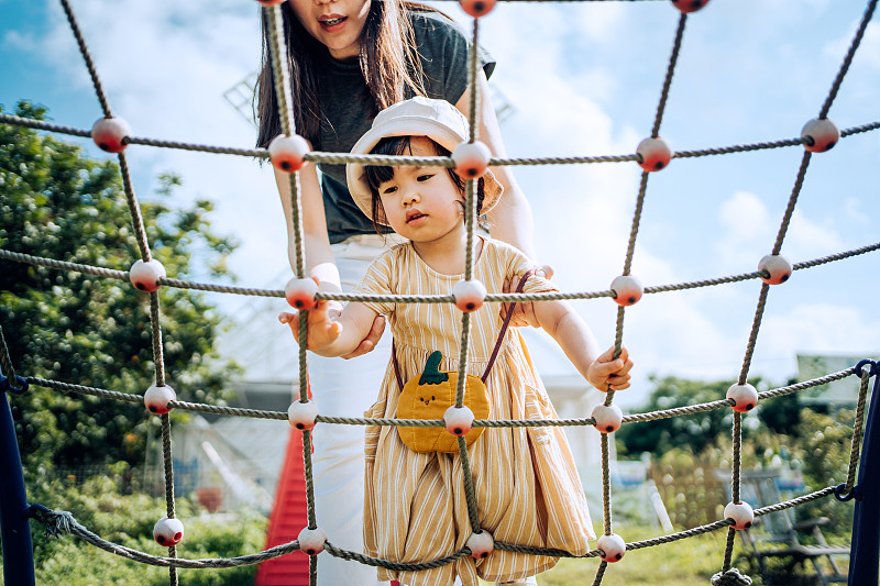 在一个阳光明媚的日子，一位年轻的亚洲母亲在户外操场上帮助小女儿攀爬绳索图片素材