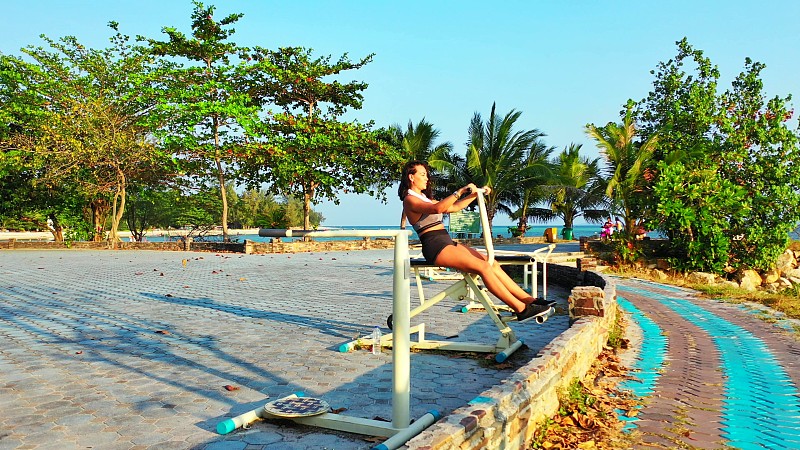 快乐的女孩在户外健身工具在美丽的度假花园周围的热带树木附近的蓝色大海图片素材