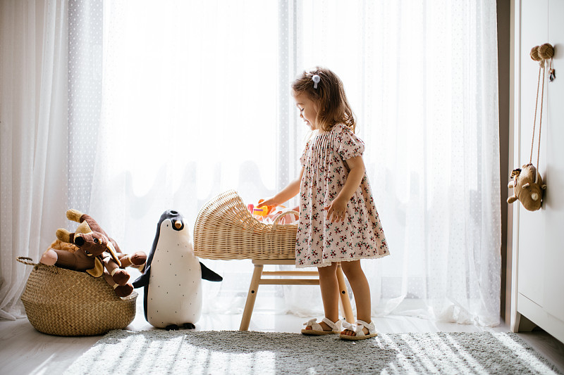 可爱的小女孩在婴儿床里喂她的玩具娃娃，在有大窗户的阳光卧室里玩图片素材