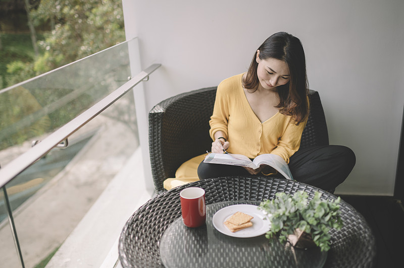 一位亚洲华裔美女中午在她的阳台上看书，放松，享受她的数字排毒时间，喝着咖啡，吃着饼干当点心图片下载