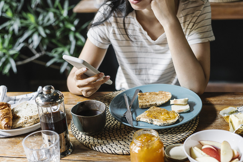 一个匿名女人使用智能手机在家里吃早餐的特写图片下载
