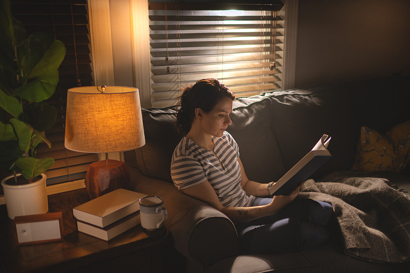 一个年轻女子坐在舒适的沙发上，在昏暗的光线下看书。图片下载