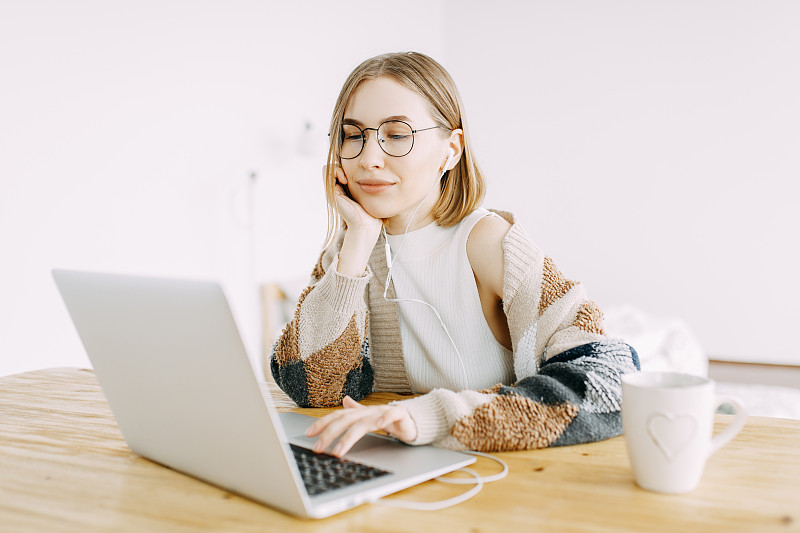 千禧一代自由职业者女孩在笔记本电脑前工作，戴着耳机听音乐，在家喝茶图片下载