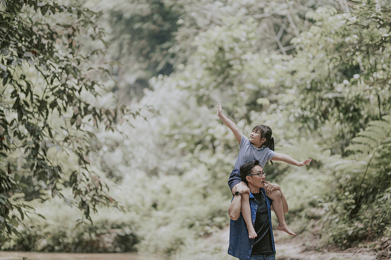 一位亚裔华人父亲在丛林中背着女儿，周末闲暇时在河边享受亲密时光图片下载