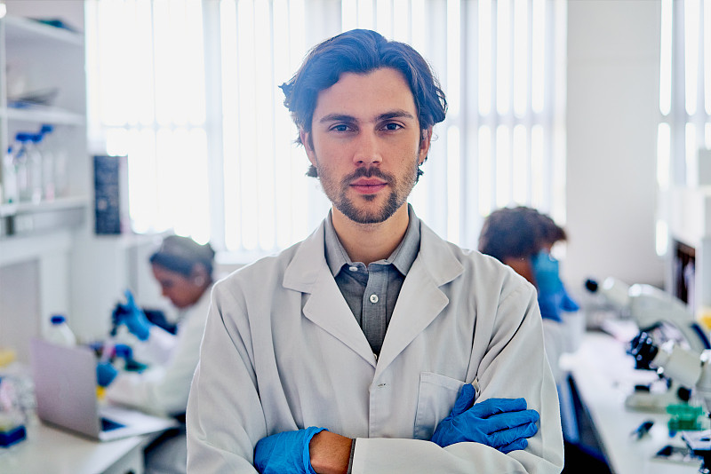 一位年轻的科学家站在实验室里，身后还有同事在工作图片下载