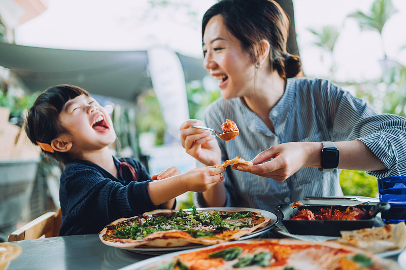 快乐的年轻亚洲母亲和可爱的小女儿在一家户外餐厅享受披萨午餐，而母亲为女儿提供肉丸和肉酱。家人享受亲密的时间和快乐的一餐在一起。家庭和外出就餐的生活方式图片下载