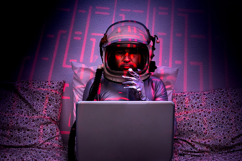 宇航员坐在沙发上，拿着笔记本电脑，凝视着无限，墙壁上有蓝色和粉色的反光，周围是蓝色的环境图片素材