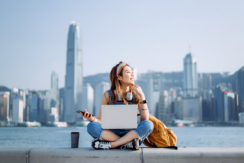 快乐的年轻亚洲女子盘腿坐在长廊，对着城市的天际线。她脖子上戴着耳机，用智能手机和笔记本电脑工作，身边放着咖啡杯。微笑着看向别处。生活方式与科技图片下载
