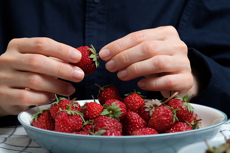 成熟的红色草莓放在一个碗或陶瓷盘子里，放在木制背景或桌子上。一个女人或一个女孩手里有一个大浆果，她会吃掉它。素食的概念，纯素和生食的营养和饮食。从农场到餐桌。蔬菜食品。摄影图片