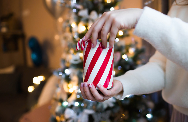 特写女人的手拿着圣诞盒和棒棒糖。闪烁的圣诞树背景上，一个女人手中的糖果。景深小，强调手和指甲。图片下载