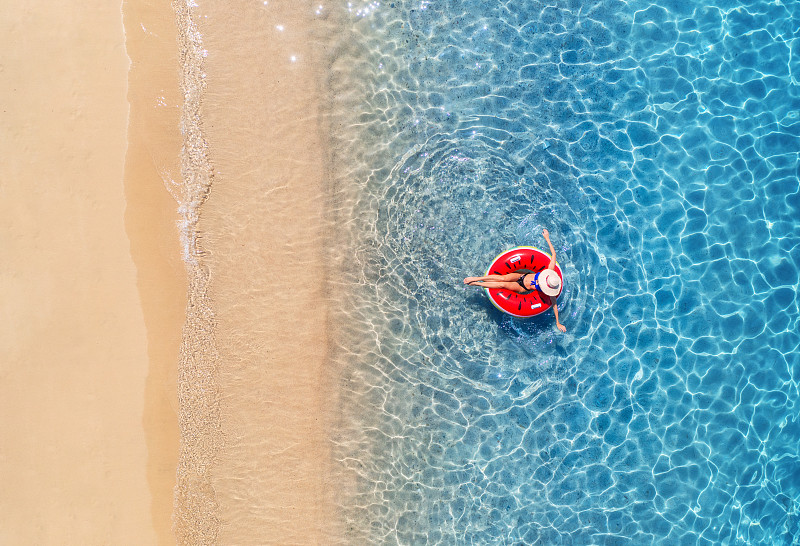 夏日日出时，一名戴着红色游泳环的帽子的女子在蓝色大海中游泳的鸟瞰图。热带风景与女孩，清澈的水，海浪，沙滩。前视图。假期。意大利撒丁岛图片下载