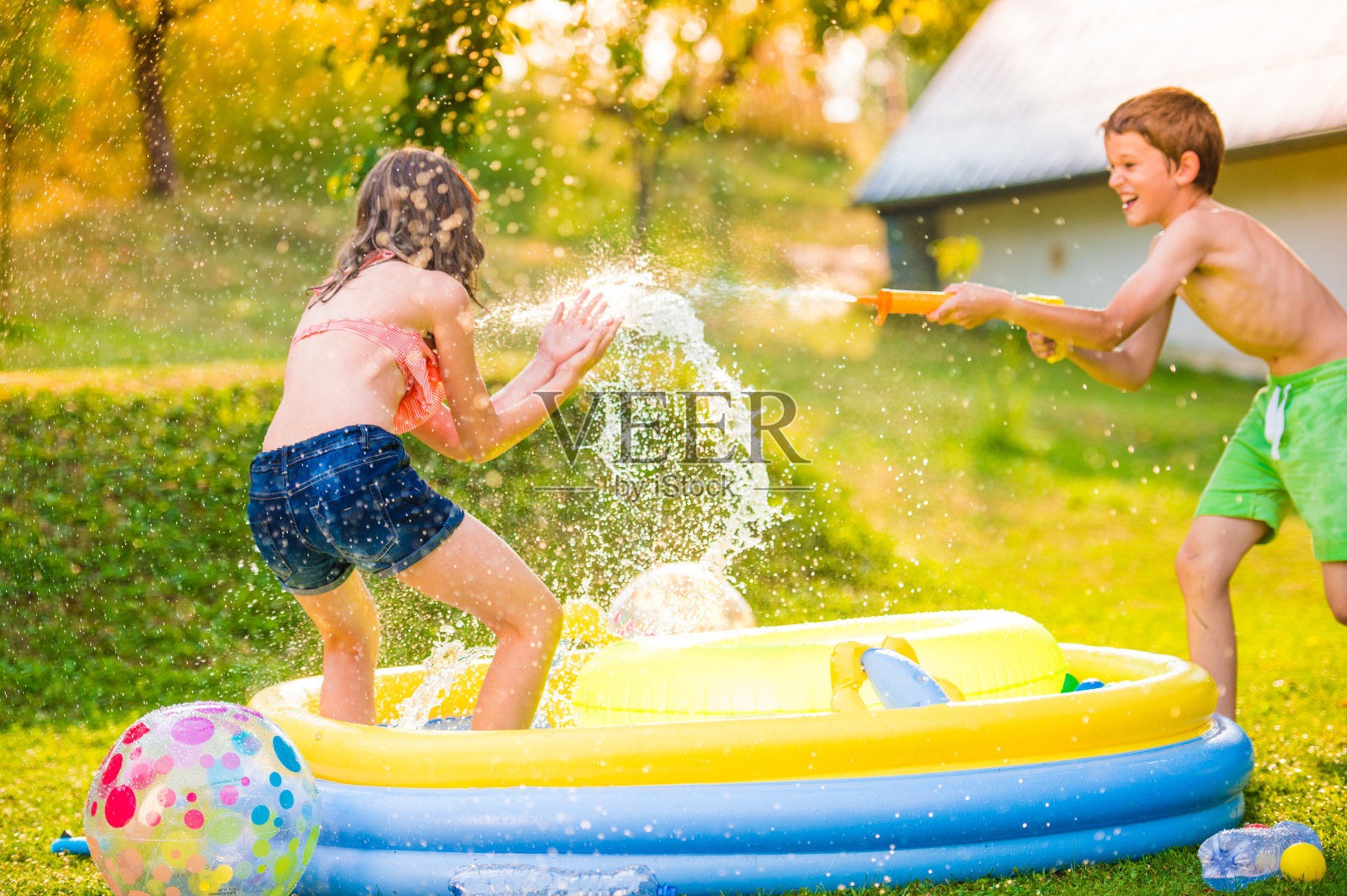 花园游泳池里，男孩用水枪向女孩泼水照片摄影图片