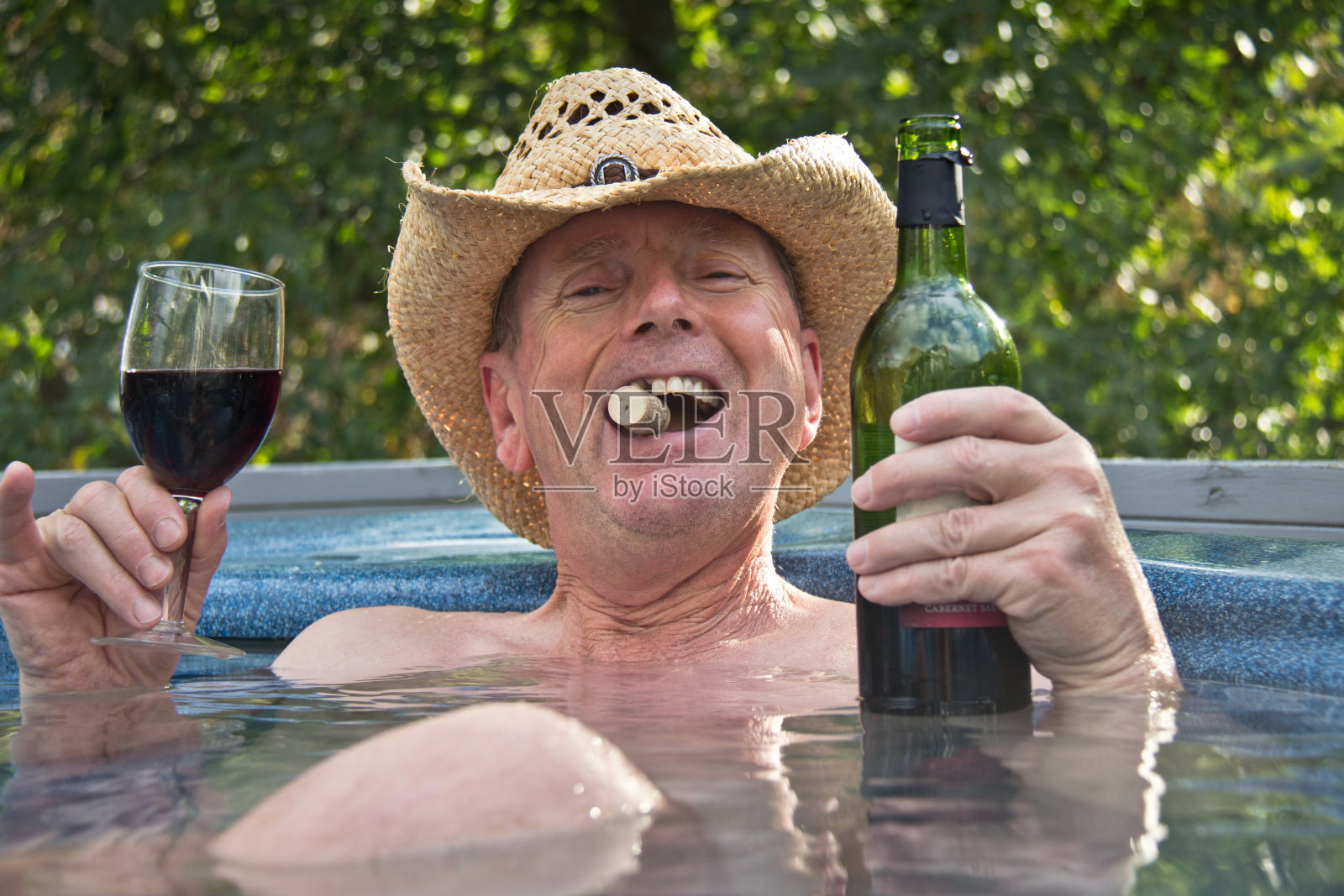 戴着牛仔帽的男人坐在热浴盆里喝着酒。照片摄影图片