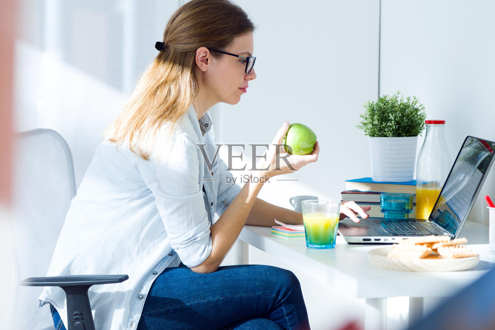 年轻漂亮的女人一边吃苹果，一边在家工作。照片摄影图片