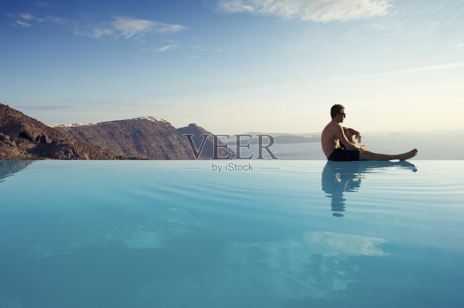 一名年轻人坐在希腊圣托里尼火山口的无边泳池边照片摄影图片