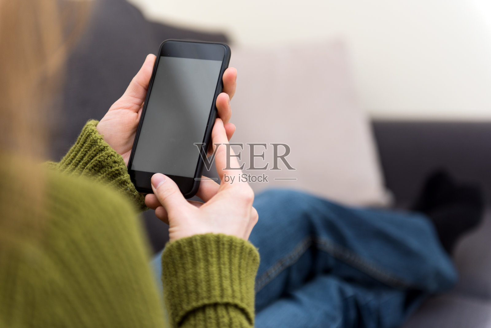 一个金发的年轻女子正坐在家里，手里拿着手机，看着一个空白的智能手机。照片摄影图片