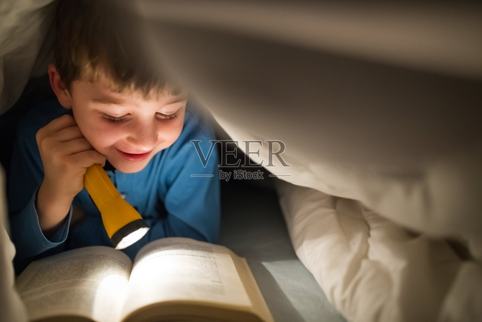 小男孩用手电筒在被子里看书照片摄影图片