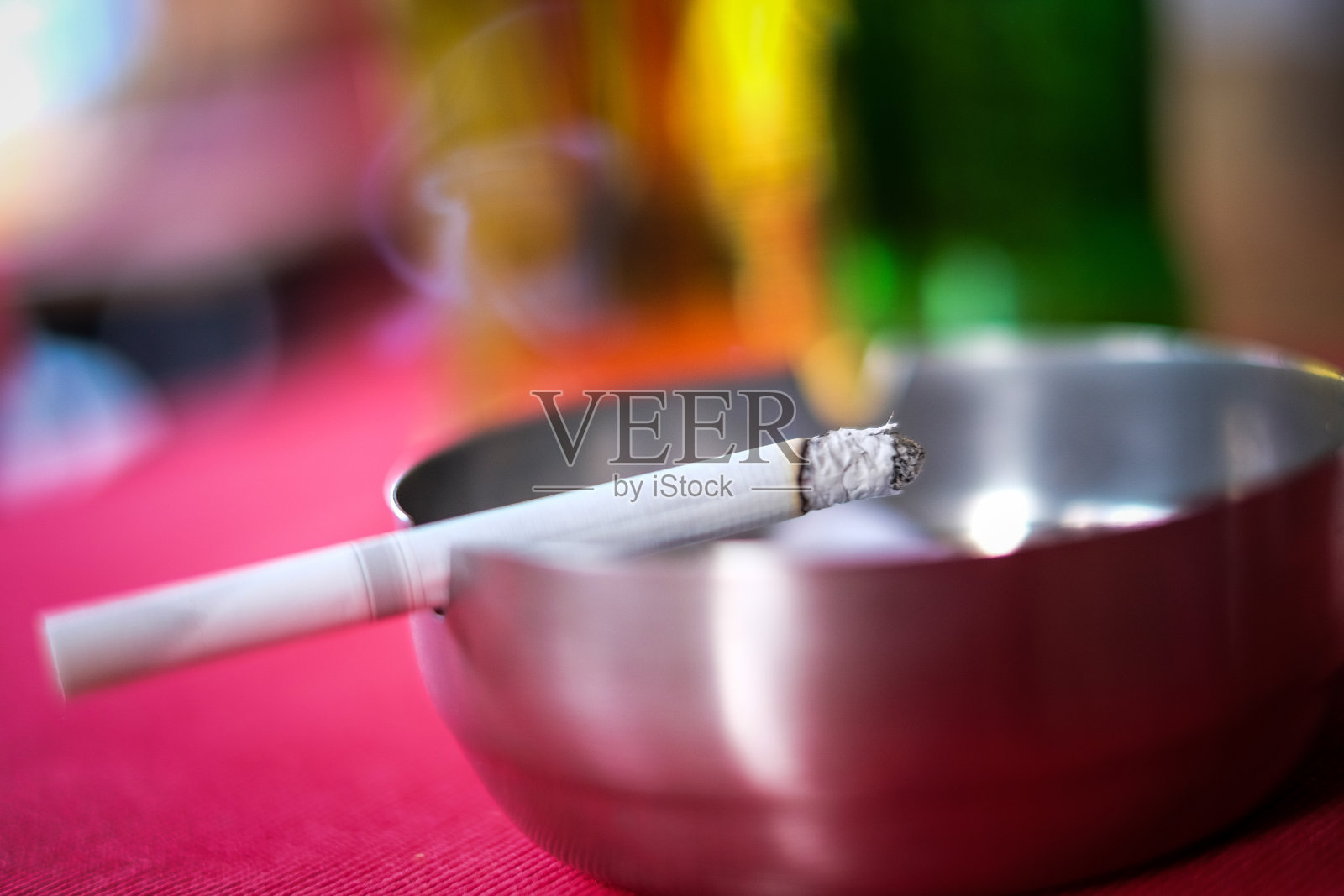 一个点燃的香烟休息在一个银色的烟灰缸与啤酒在背景的特写照片摄影图片