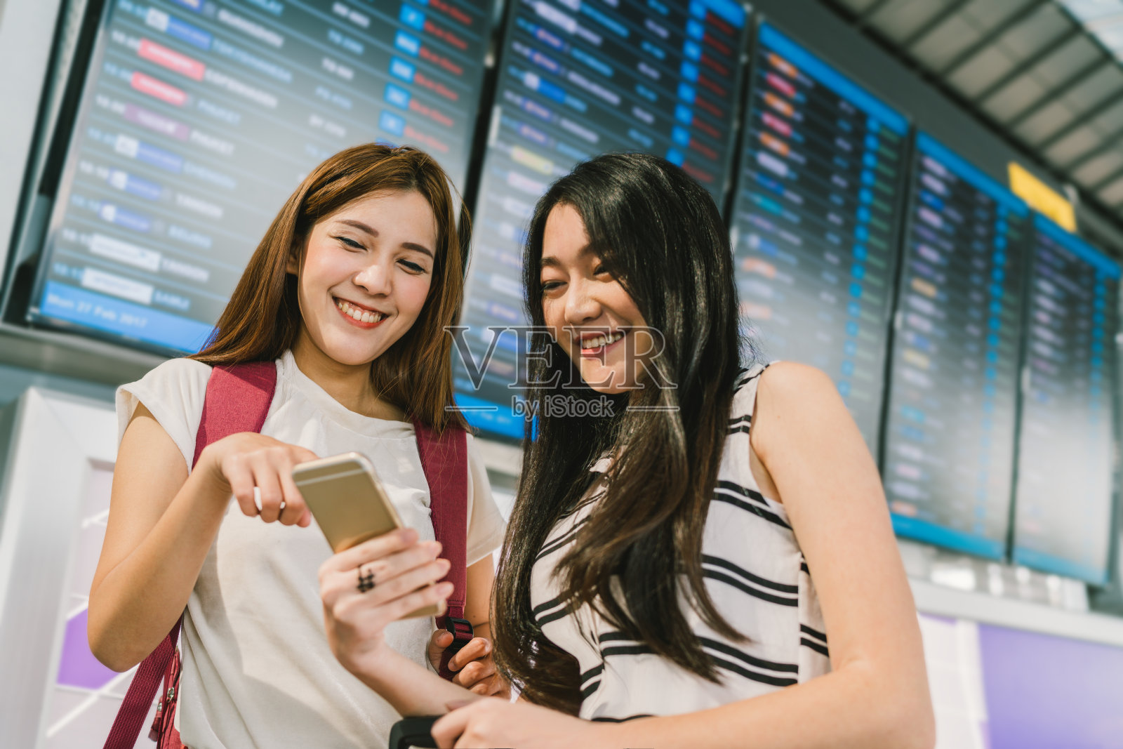 两个亚洲女孩在机场的航班信息板上一起使用智能手机。在线登记，时间表申请，或假日旅游概念照片摄影图片