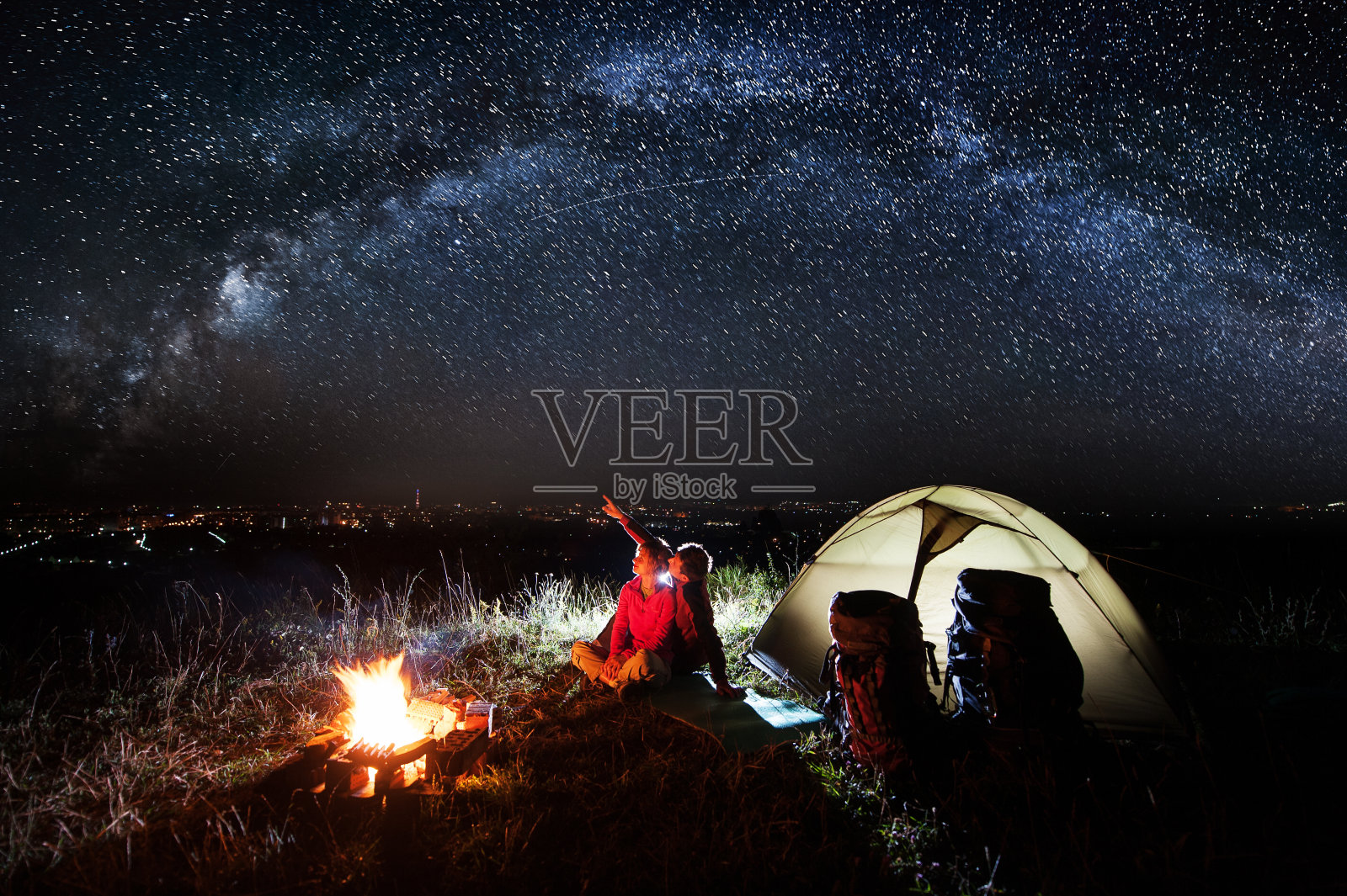 晚上在镇上附近露营。年轻的夫妇坐在篝火和帐篷附近，看着美丽的星空，欣赏夜景。盖伊指着天空。天体摄影术照片摄影图片