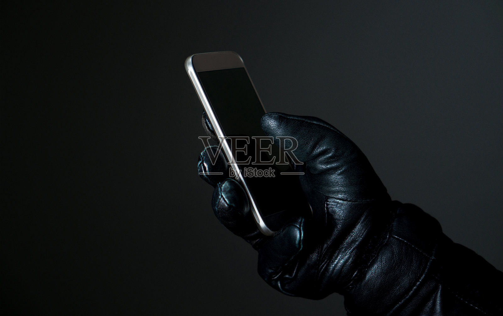 移动和网络安全线程概念。黑暗中戴着黑色皮手套拿着智能手机的罪犯。黑客触摸智能手机屏幕。照片摄影图片