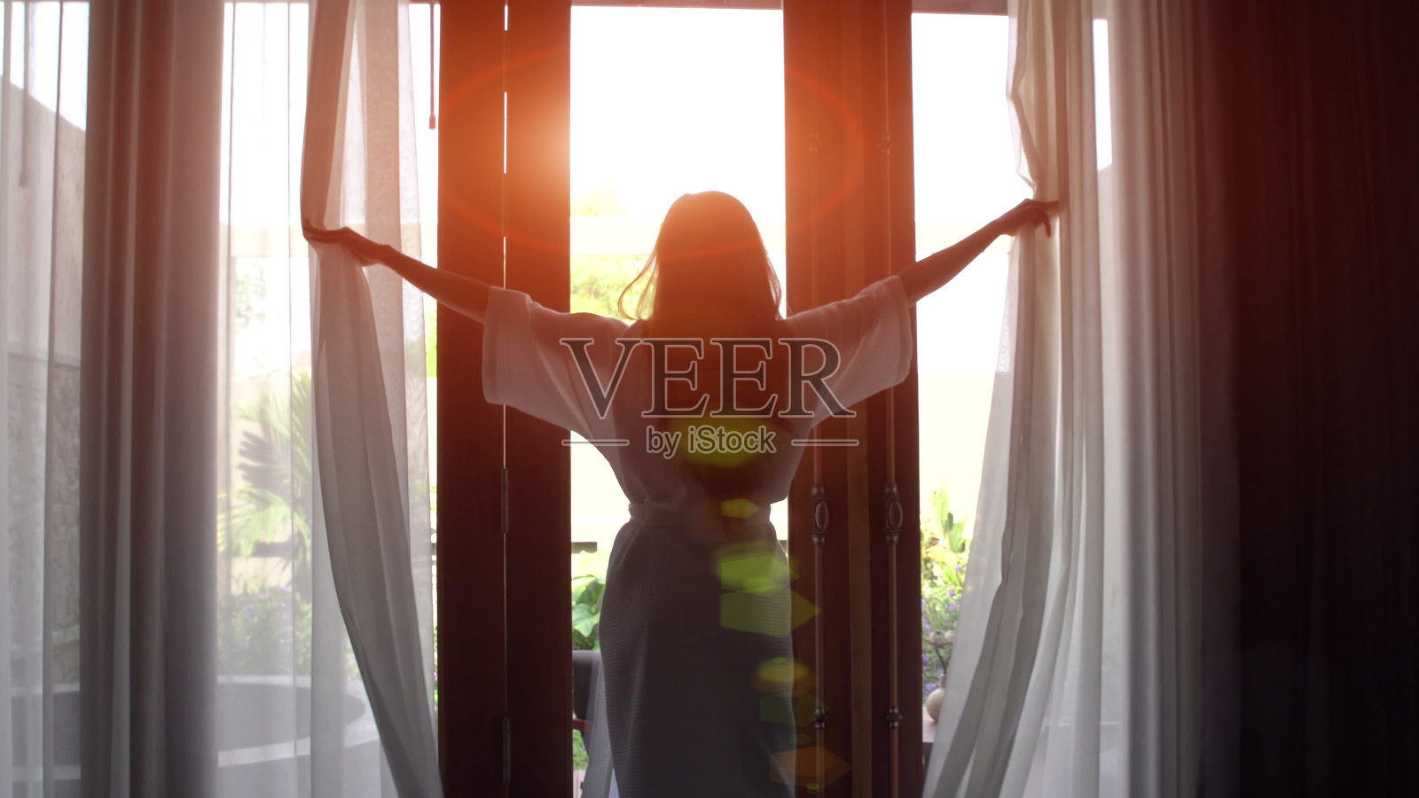 穿着浴袍的年轻女子拉开窗帘，伸懒腰，站在家里靠近窗户的地方。照片摄影图片