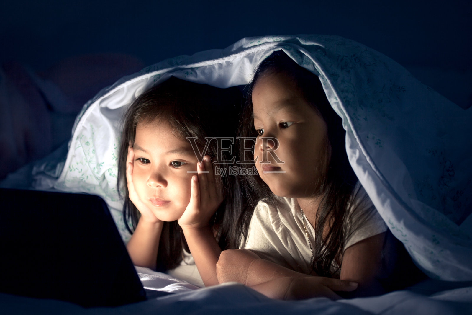 两个亚洲小女孩晚上在毯子下使用电子平板电脑照片摄影图片