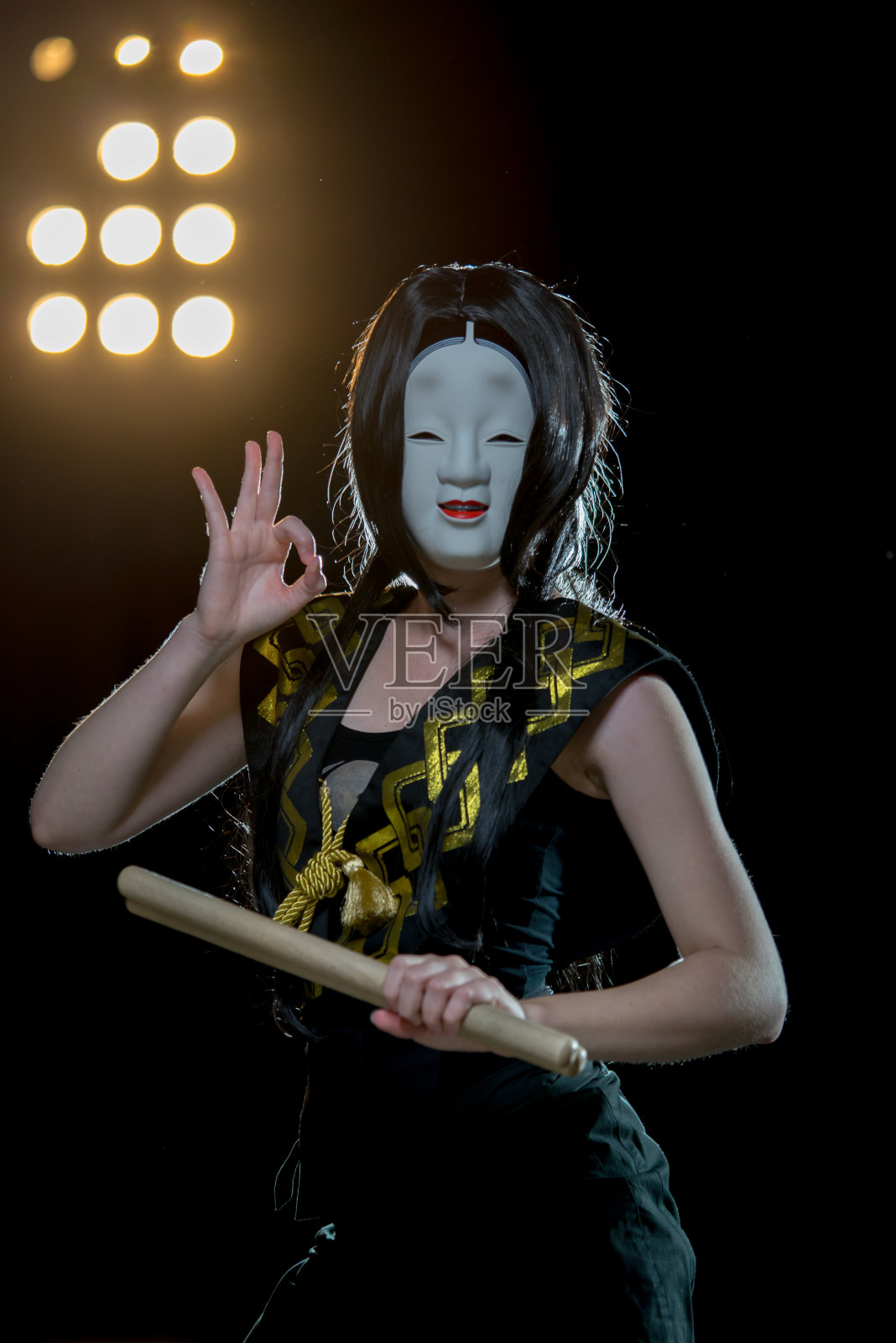 鼓手女孩在白色恶魔面具与鼓槌显示ok的视线，工作室音乐会拍摄在一个黑暗的背景和天窗灯。照片摄影图片