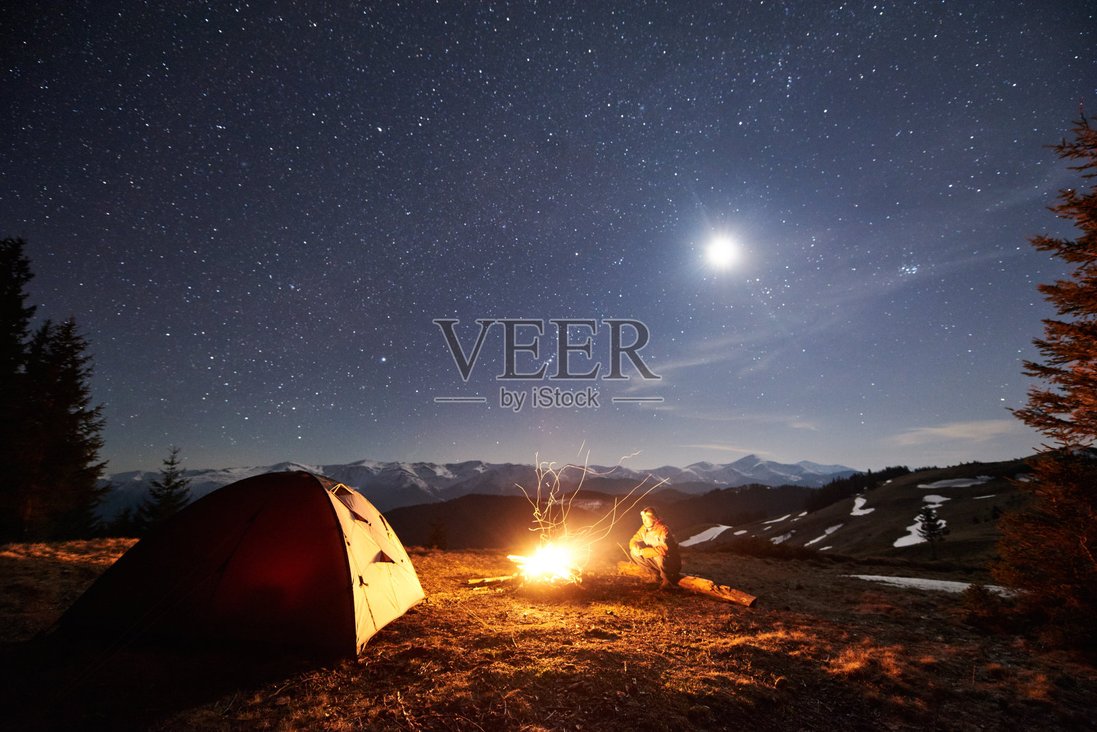 男性徒步旅行者晚上在森林附近的营地休息。在满是星星和月亮的美丽夜空下，坐在篝火和帐篷旁的人，在山上欣赏夜景照片摄影图片